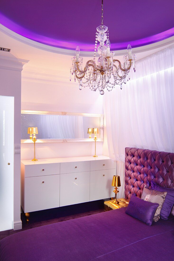 Elegantes Schlafzimmer in Violetttönen und goldfarbenen Tischleuchten auf weißer Schminkkommode, lilafarbene Deckengestaltung und Kristallkronleuchter