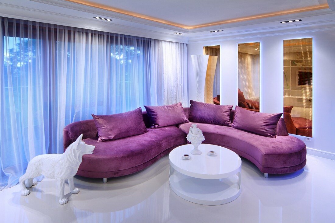 Lila Polstersofa und weißer, runder Designer-Couchtisch in offenem Wohnraum mit stimmungsvoller Beleuchtung
