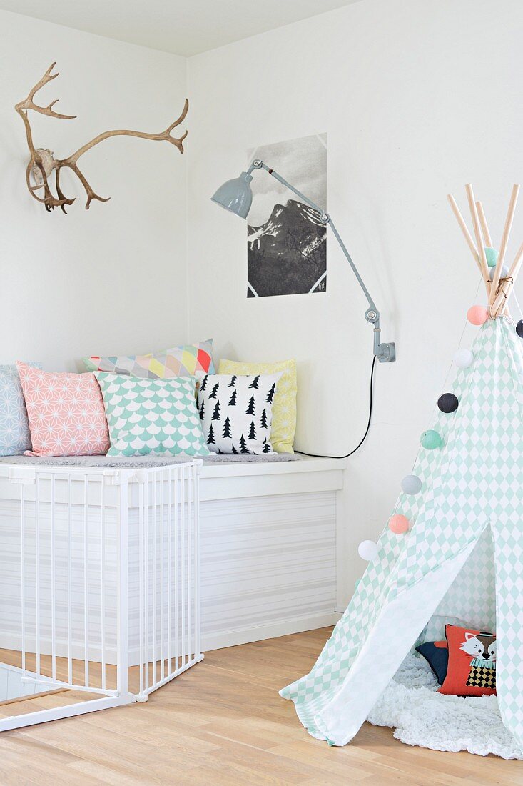 Indoor teepee in pastel shades next to cosy seating area with scatter cushions below stag's antlers and picture