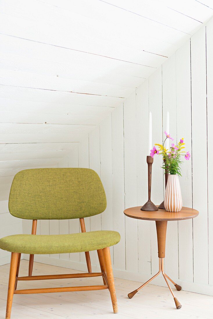 Retro upholstered chair and candles and flowers on side table in white, wood-clad attic room