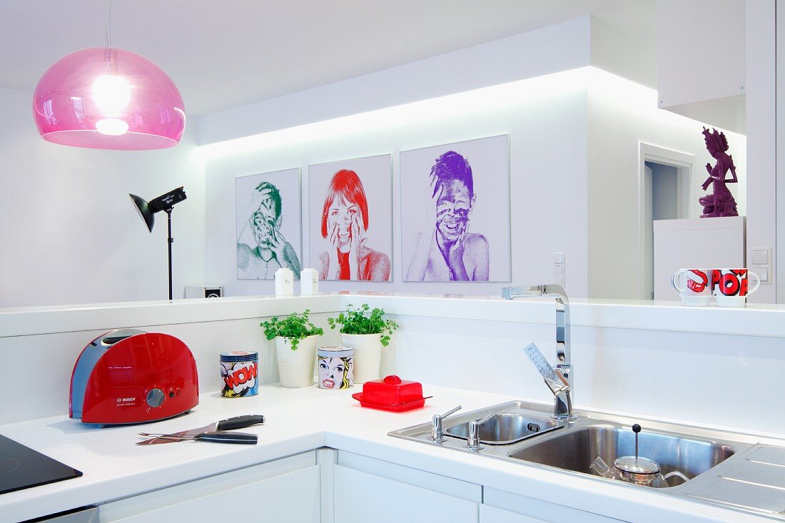 Blick über weiße Küchentheke auf Portraitfotografien an weißer Wand und rosafarbene Pendelleuchte