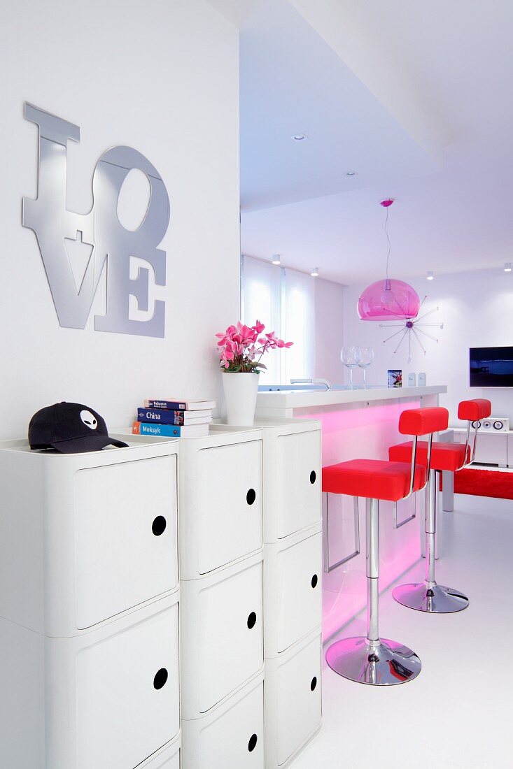 Lettering spelling 'LOVE' above sideboard and red bar stools at white kitchen counter in open-plan interior