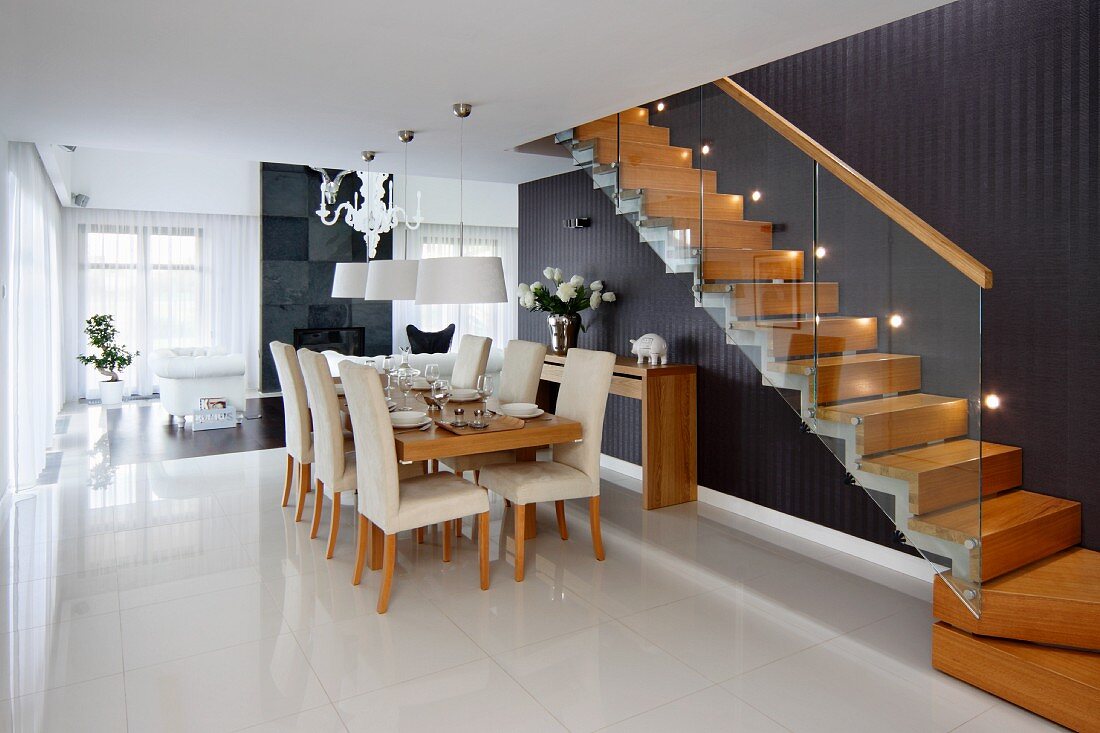 Dining area with pale, upholstered chairs below pendant lamps next to wooden staircase with glass balustrade and black wall with recessed spotlights
