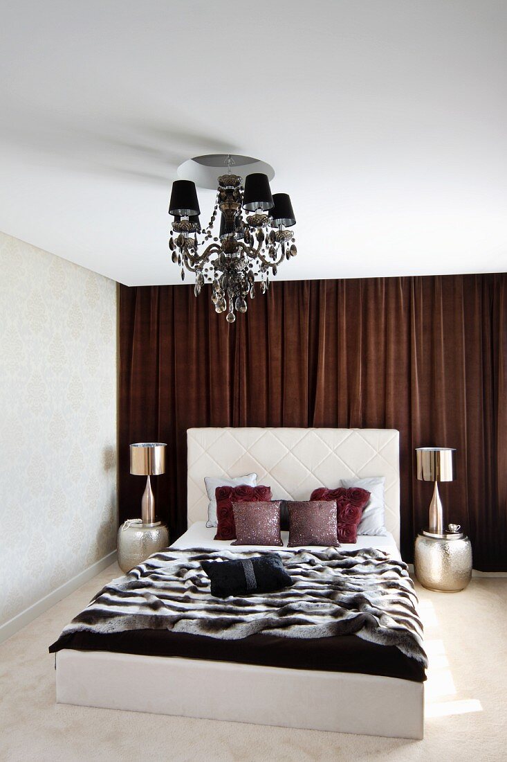Doppelbett mit hohem Kopfteil und Felldecke, seitlich orientalische Metall Beistelltische und Tischleuchten, vor braunem Vorhang