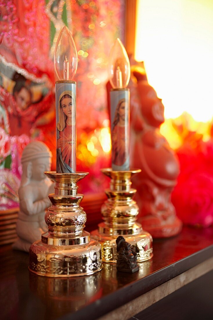 Elektrische Kerzenhalter mit christlichen Bildmotiven, winzige Buddhafigur und indische Figuren im Hintergrund auf einer Ablage