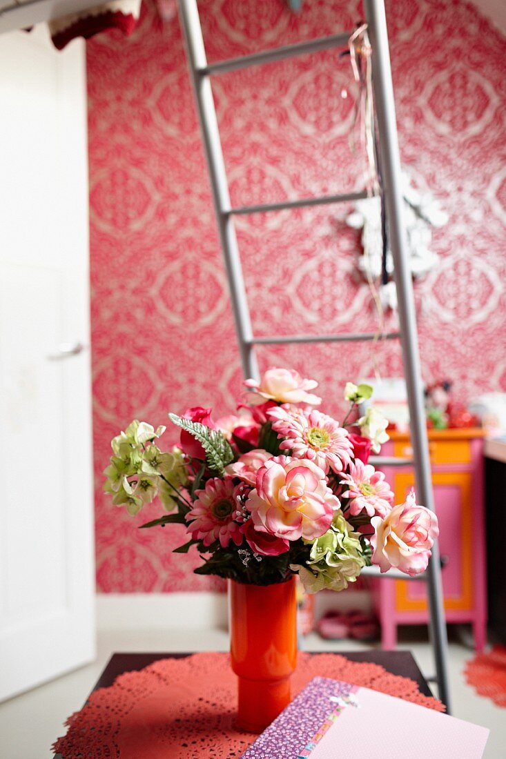 Blumenstrauss in orangefarbener Glasvase auf Spitzendeckchen, dahinter eine Metallleiter und pinkfarbenes Tapetenmuster