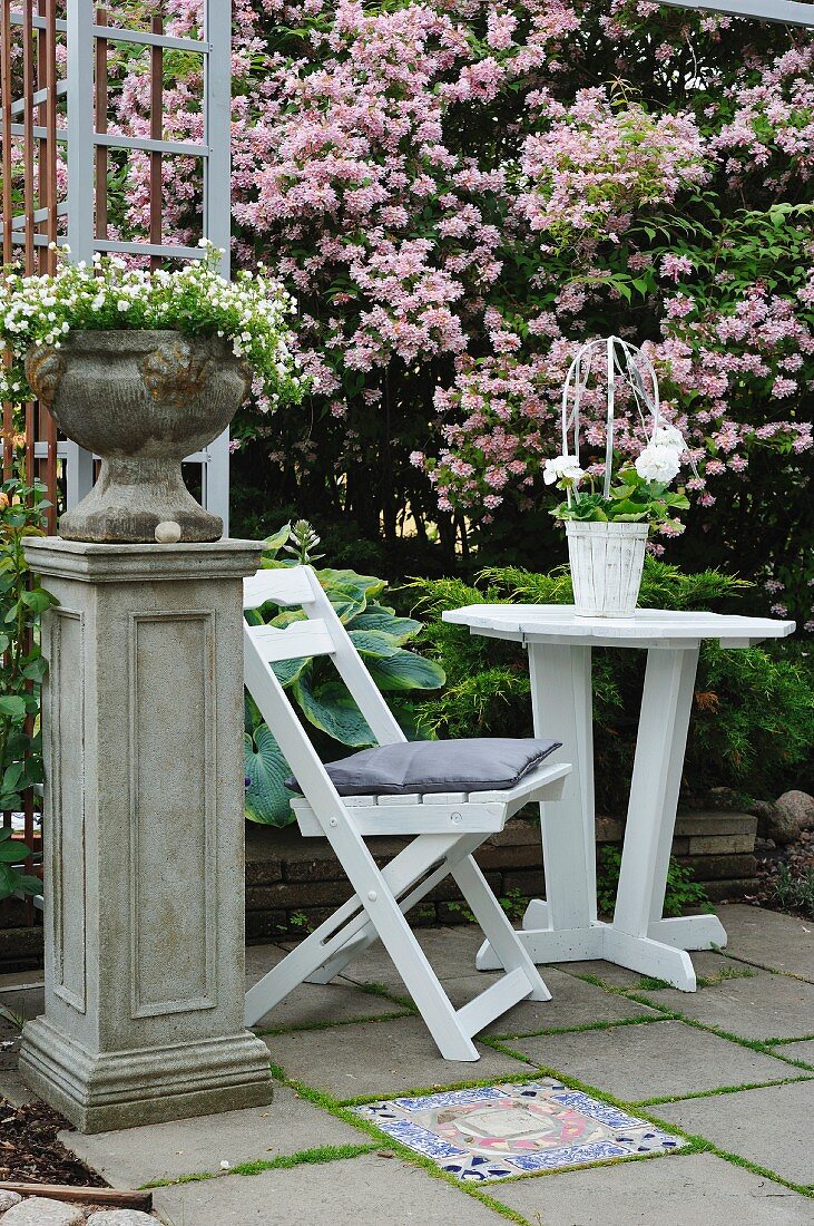 Bepflanzte Amphore auf Sockel neben weißem Gartenstuhl mit Tisch auf Terrasse