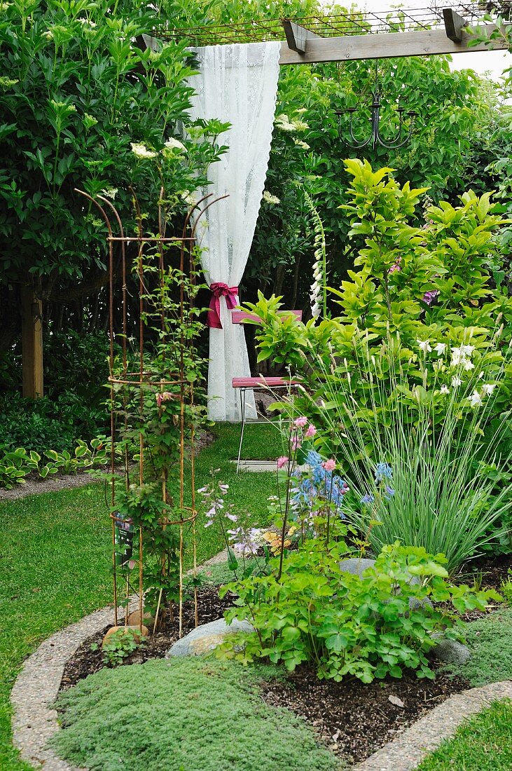Blumenbeet im Garten, im Hintergrund geraffter weißer Spitzenvorhang an Pergola aufgehängt