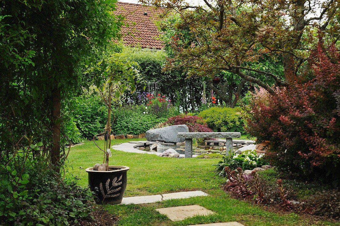 Sommerlicher Garten mit Bäumchen im Topf, Steinbank und Findling am runden Gartenteich