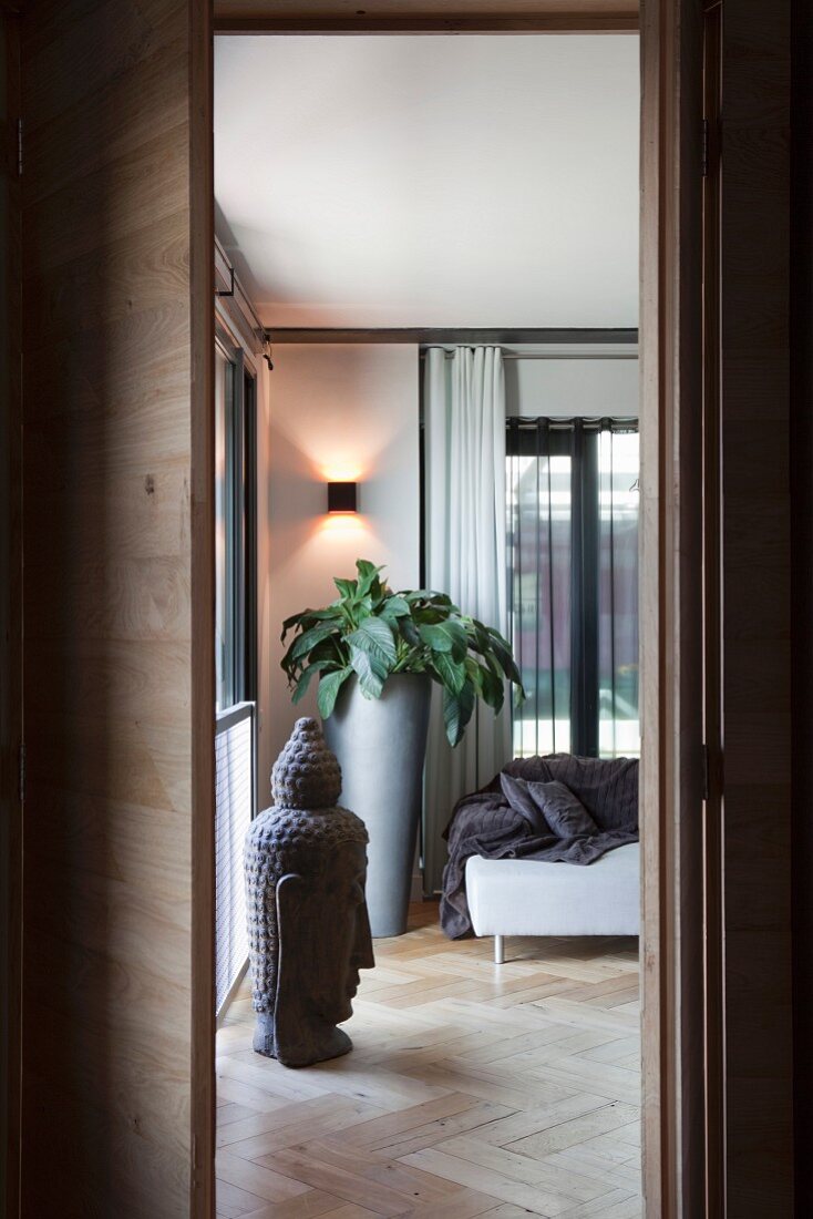 Blick durch offene Tür, Buddhabüste auf Fischgrätparkett und hohes graues Pflanzgefäss im Zimmerecke