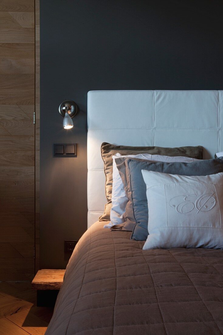 Mehrere Kissen auf Bett mit weißem Polsterkopfteil an dunkelgrauer Schlafzimmerwand mit Wandleuchte
