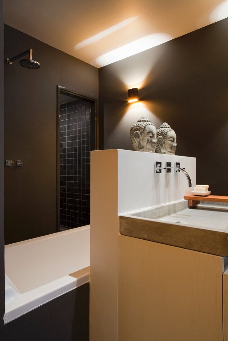Designer washstand against masonry partition with two heads of Buddha on top screening bathtub and shower area in dark brown bathroom