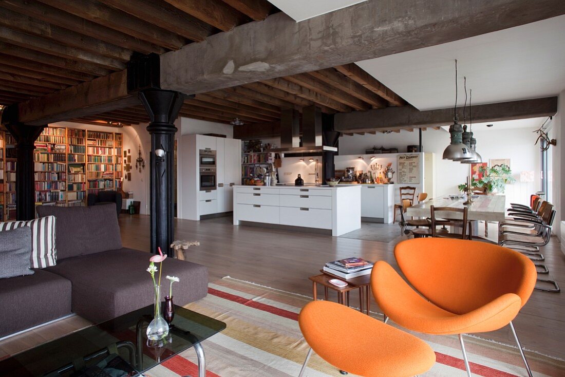 Orangefarbener Retro-Sessel mit passendem Fussschemel im Designerstil in Loungebereich, offene Küche und Essplatz im Loft