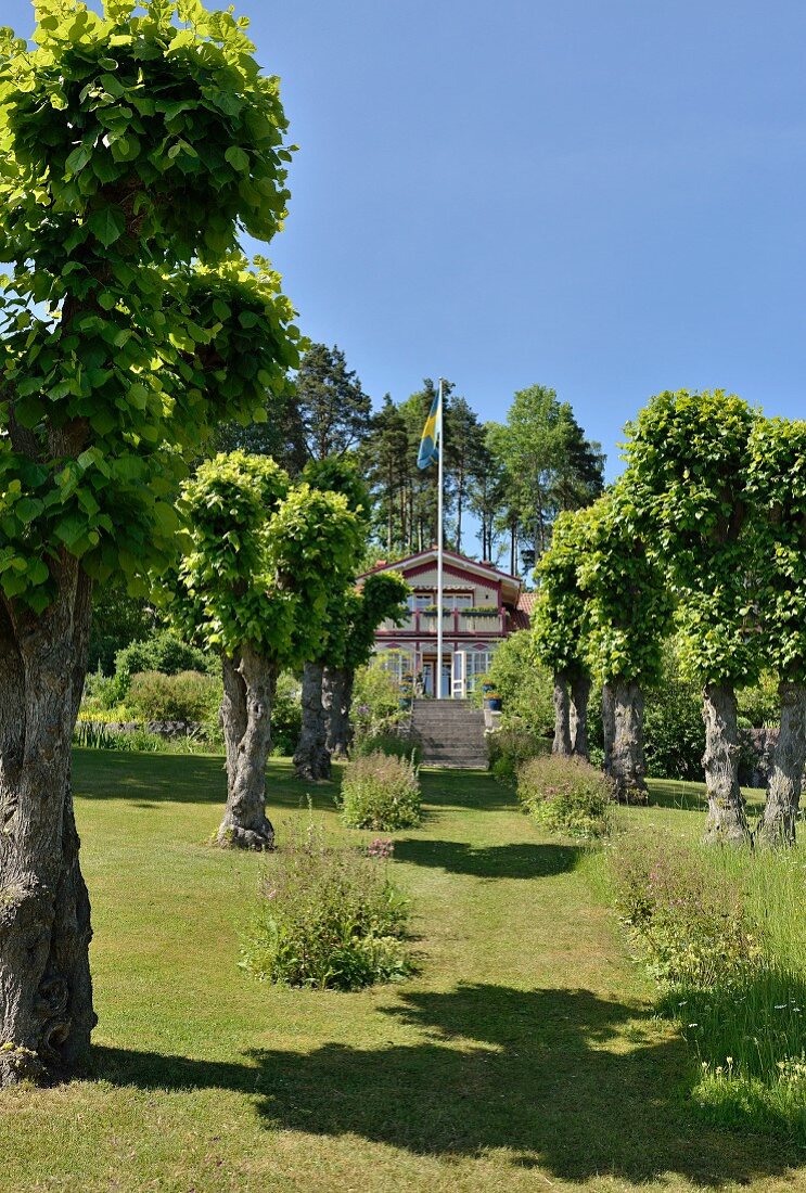 Sommerlicher Garten mit Allee zum Landhaus mit schwedischer Fahne und Treppenaufgang