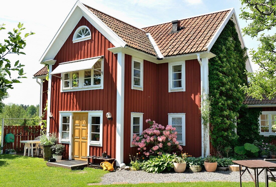 Rotbraun gestrichenes Holzhaus mit weissen Fenstern, davor gekieste Fläche mit Blumentöpfen und Azalea