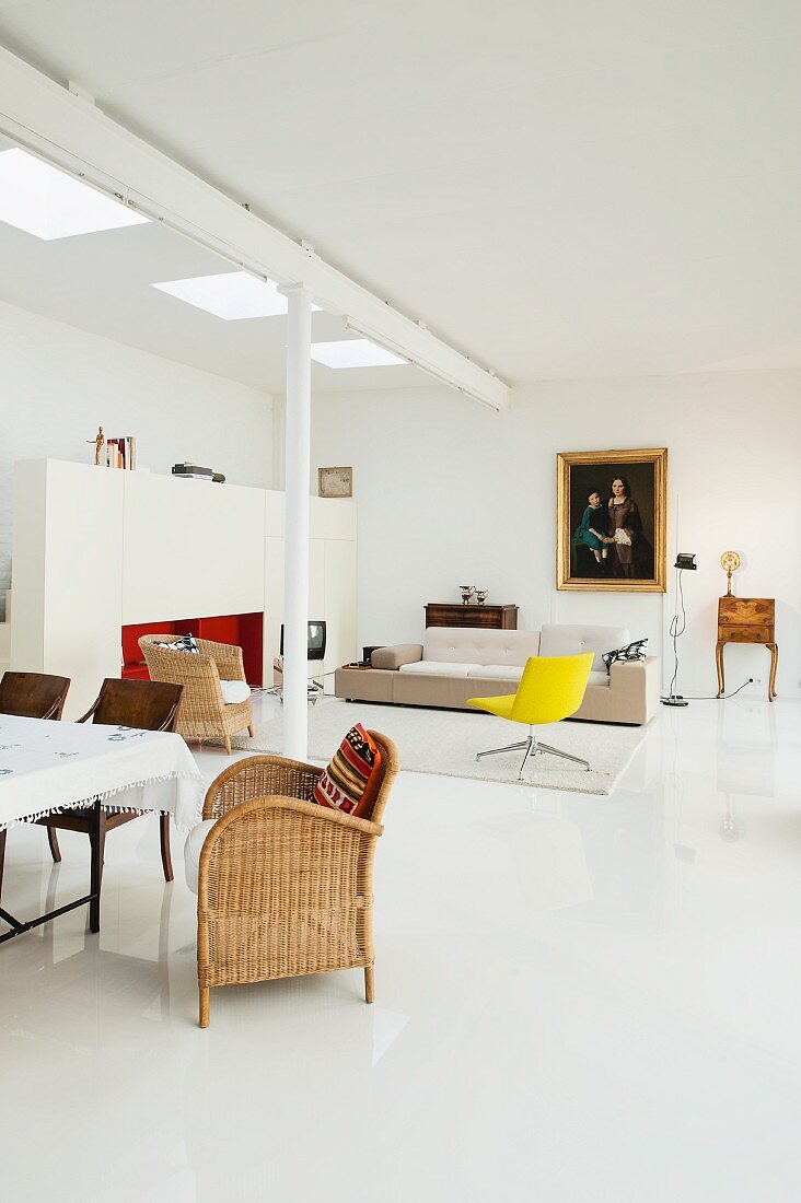 Minimalistische Loftwohnung, im Vordergrund Rattanstuhl vor Essplatz, im Hintergrund Loungebereich, gelber Drehsessel und Designersofa