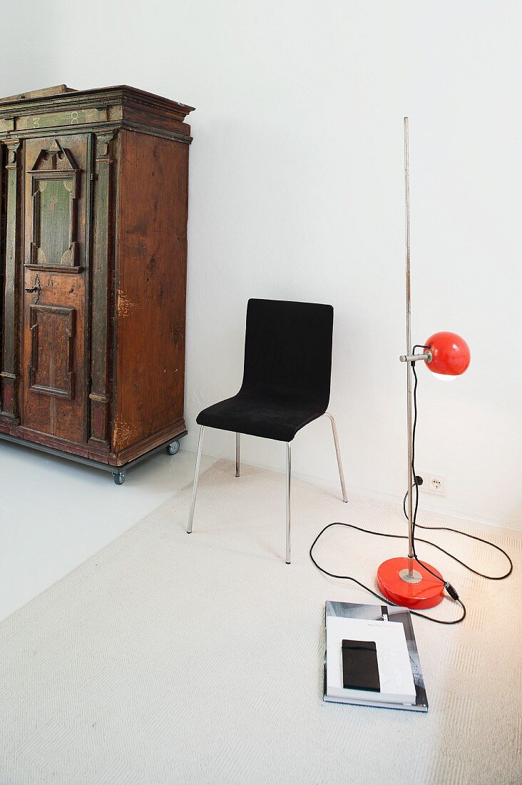 Retro standard lamp with red, spherical lampshade and black chair next to antique cupboard against white wall