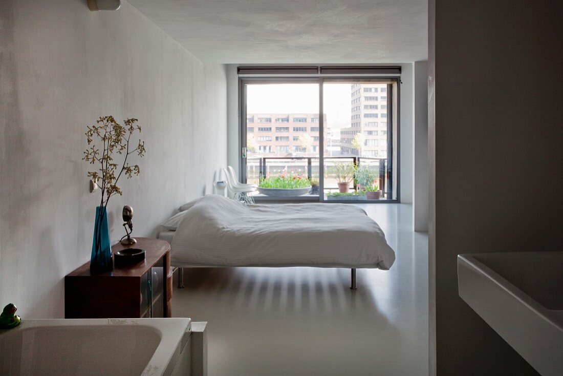 Blick vom Bad Ensuite in minimalistisches Schlafzimmer mit Doppelbett, im Hintergrund Glastür zu Balkon