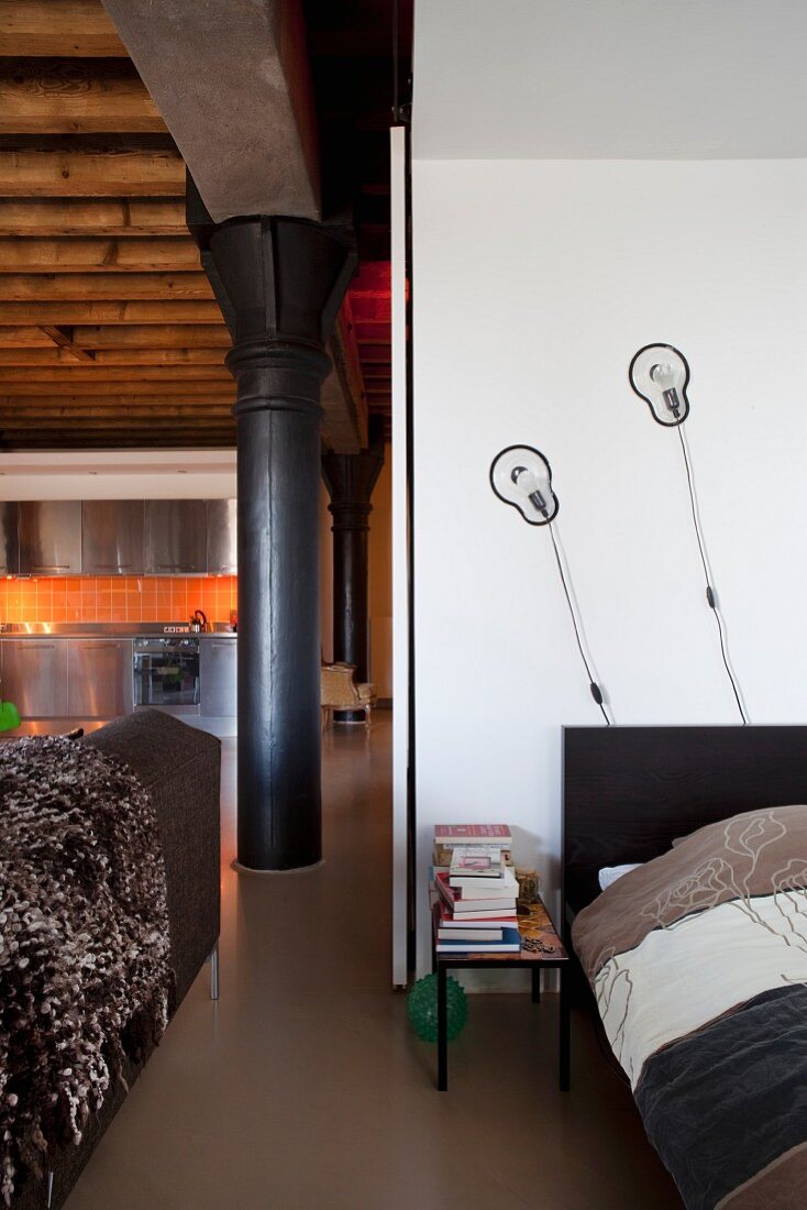 Offener Schlafbereich mit Designerwandleuchten, Blick auf schwarze Metallsäulen und Edelstahlküche in Loftwohnung