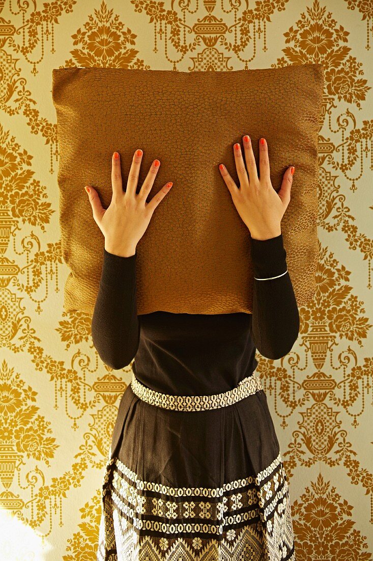 Frau mit lackierten Fingernägeln hält Kissen vor Gesicht, dahinter goldene Ornament Tapete an Wand