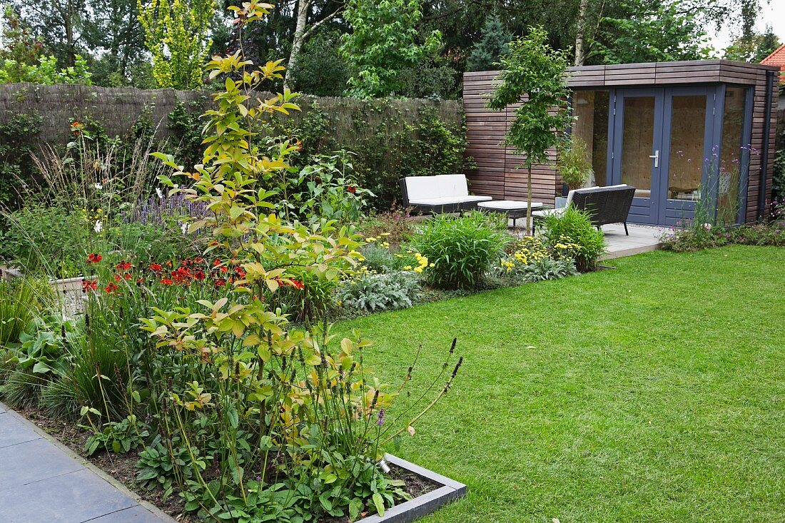 Beds of flowering plants in garden with seating area on terrace and summer house