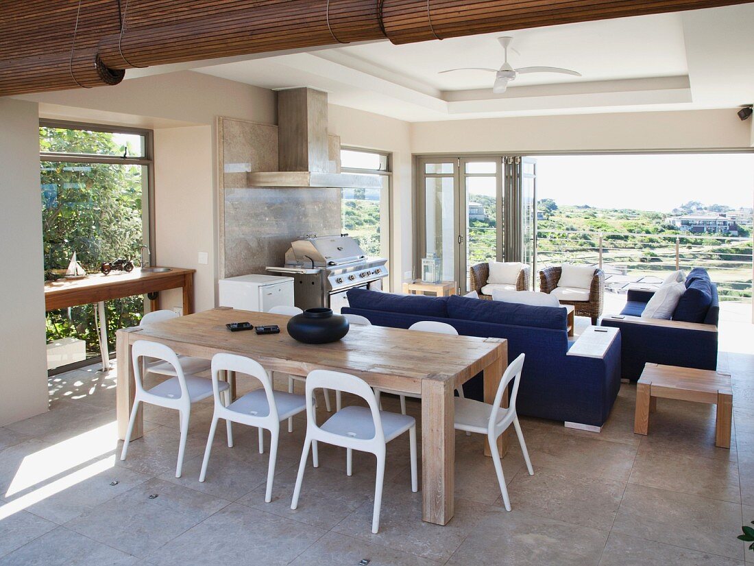 Solid wooden table, white plastic chairs, blue sofas and view of countryside through open folding terrace doors