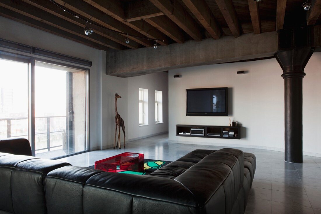 Black leather couch below wood-beamed ceiling and black metal column in industrial-style loft apartment with minimalist furnishings