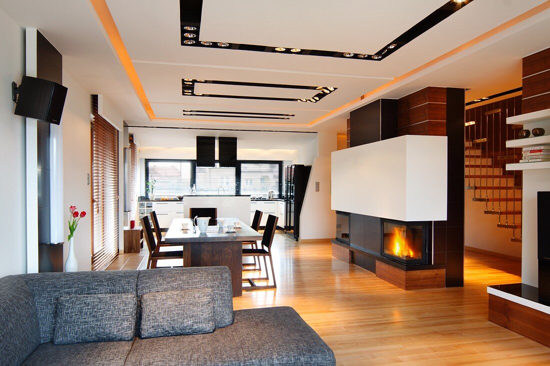 View past grey couch into loft-style apartment; dining area next to fireplace in partition and recessed spotlights in suspended ceiling