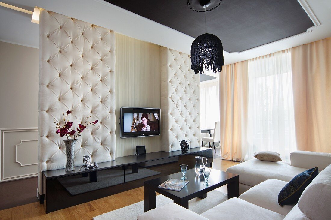 Eleganter Wohnraum mit abgehängter Decke, weisser Sofalandschaft und schwarzem Sideboard vor gepolsterter Raumteilerwand