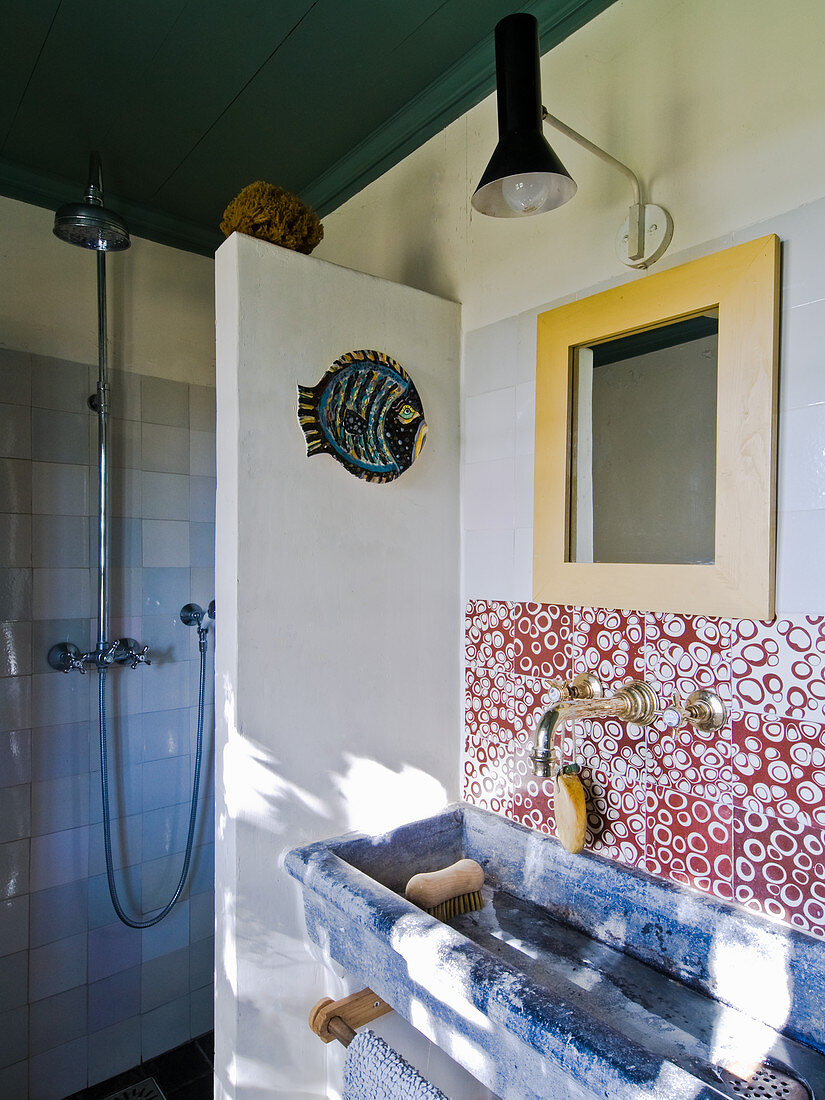 Trogartiges Waschbecken vor gefliester Wand mit rot-weißem Kringelmuster, seitlich Trennwand vor Duschbereich in ländlichem Bad