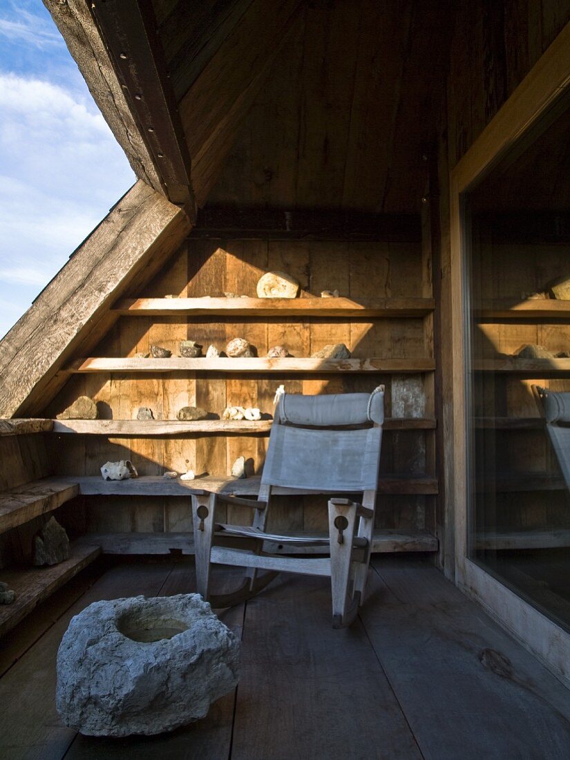 Rustikaler Sessel mit Segeltuchbezug auf Dachterrasse, vor sonnenbeschienenem Regal mit Steinsammlung, im Vordergrund Felsbrocken auf Holzdielen