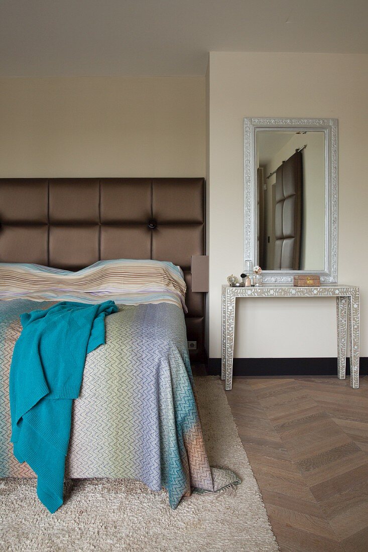 Console table below mirror next to bed with tall, brown, upholstered headboard and bedspread with subtle gradient of colours