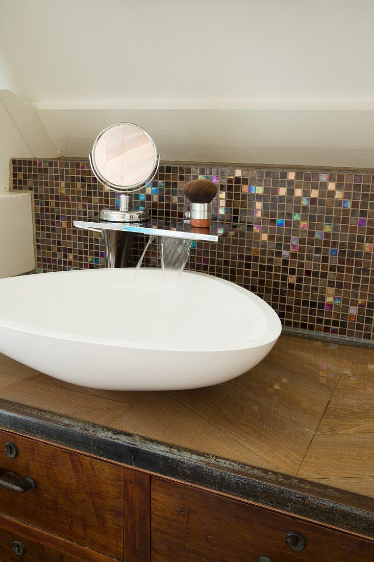 Amorphous basin on custom, wood-veneer washstand with mosaic-tiled splashback and shaving equipment on shelf