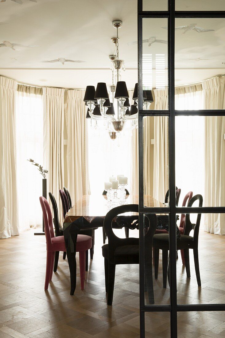 Dining area with antique chairs below pendant lamp with small lampshades; floor-length curtains on bay window in background