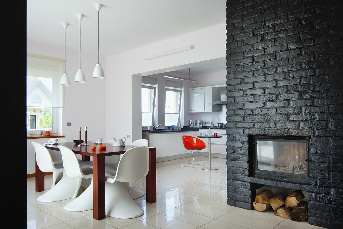 Modern, open-plan interior with retro character - white classic chairs around dining table and fireplace in black-painted brick wall to one side