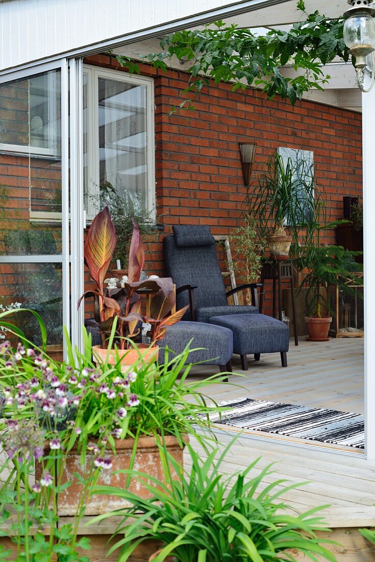 Topfpflanzen auf Terrasse, dahinter offene Schiebetür mit Blick auf Sessel mit passenden Fussschemel vor Ziegelwand
