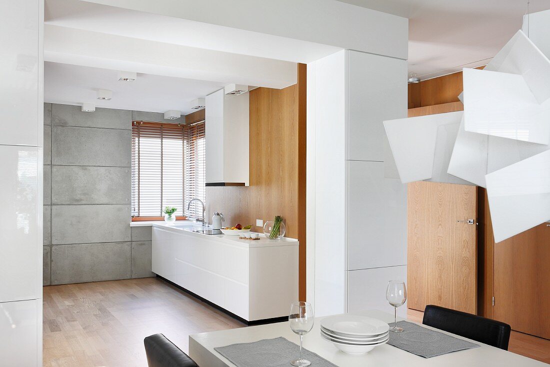 Blick über Essplatz in offene Küche mit weißem, minimalistischem Küchenblock und passendem Dunstabzug vor Holzwand