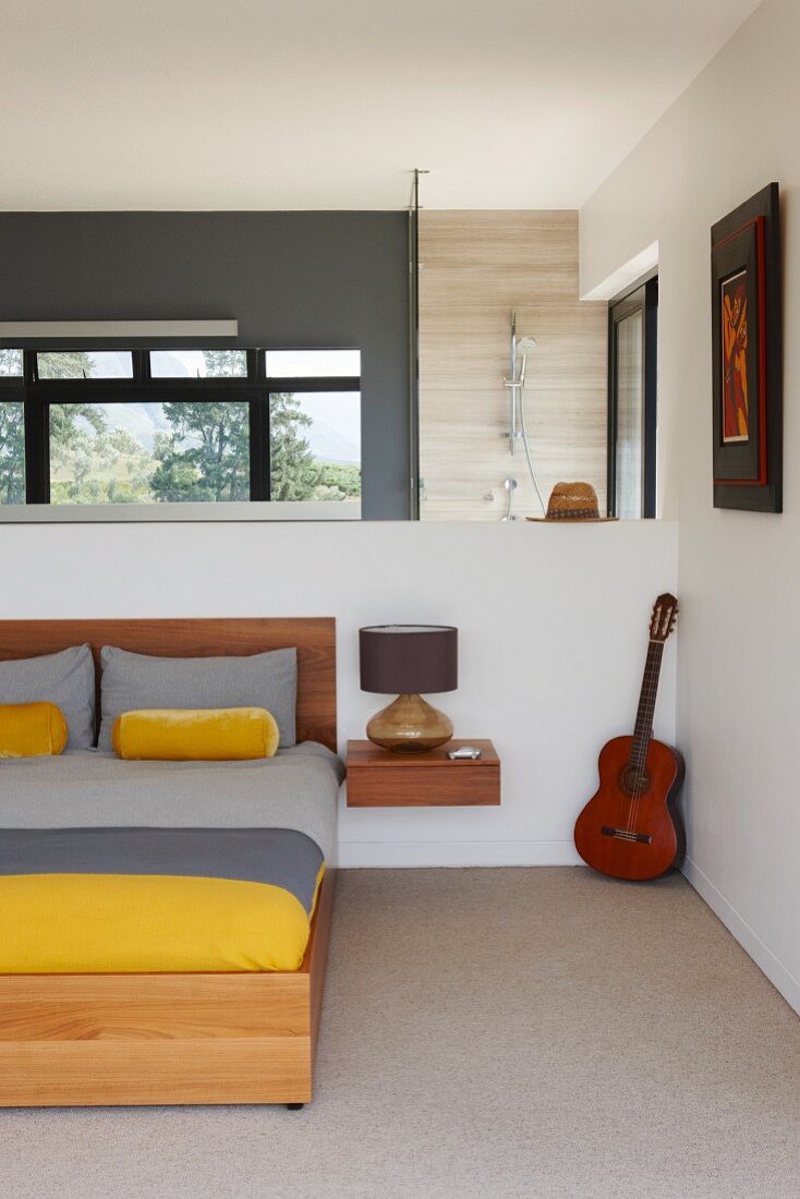 Double bed with grey and yellow bed linen and minimalist bedside cabinet on white half-height wall; ensuite bathroom in background