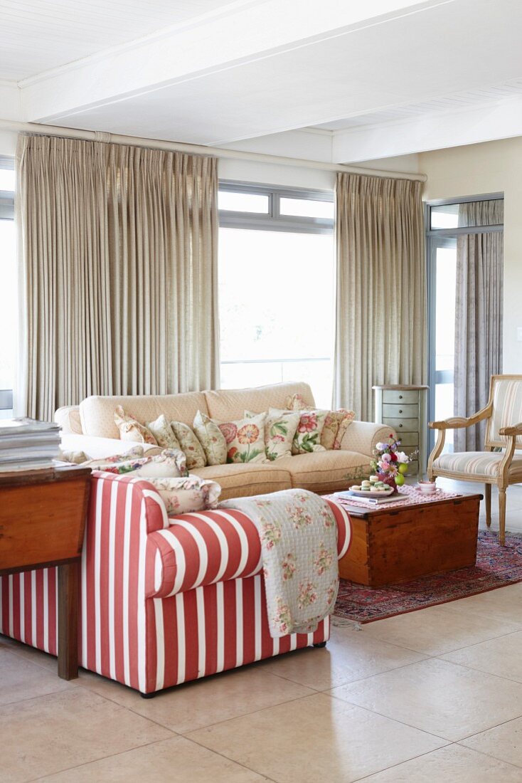 Sofa mit rot-weissen Streifen und hellem Bezug um Holztruhe, im Hintergrund bodenlange Vorhänge an Fensterfront in Wohnzimmer