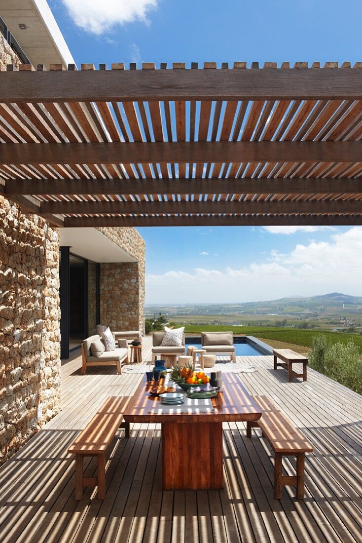 Simple modern outdoor furniture below wooden slatted pergola on spacious terrace adjoining stone house