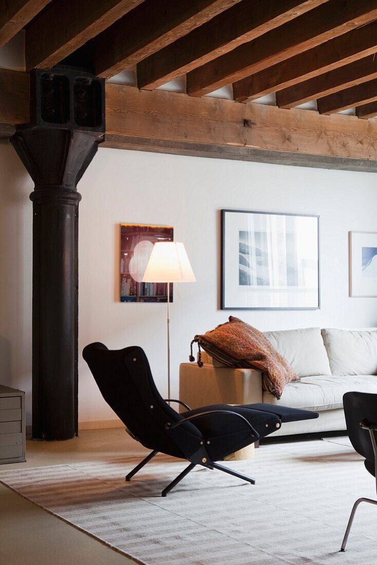 Retro-style, black armchair with footrest and standard lamp next to sofa in loft-apartment with industrial character