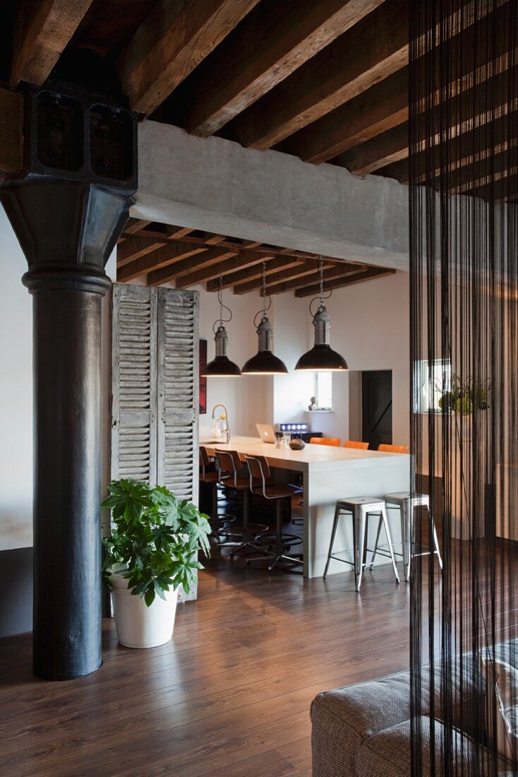 Industrial charm in loft apartment with black metal column under concrete girder and wood-beamed ceiling; modern dining area with row of pendant lamps in background