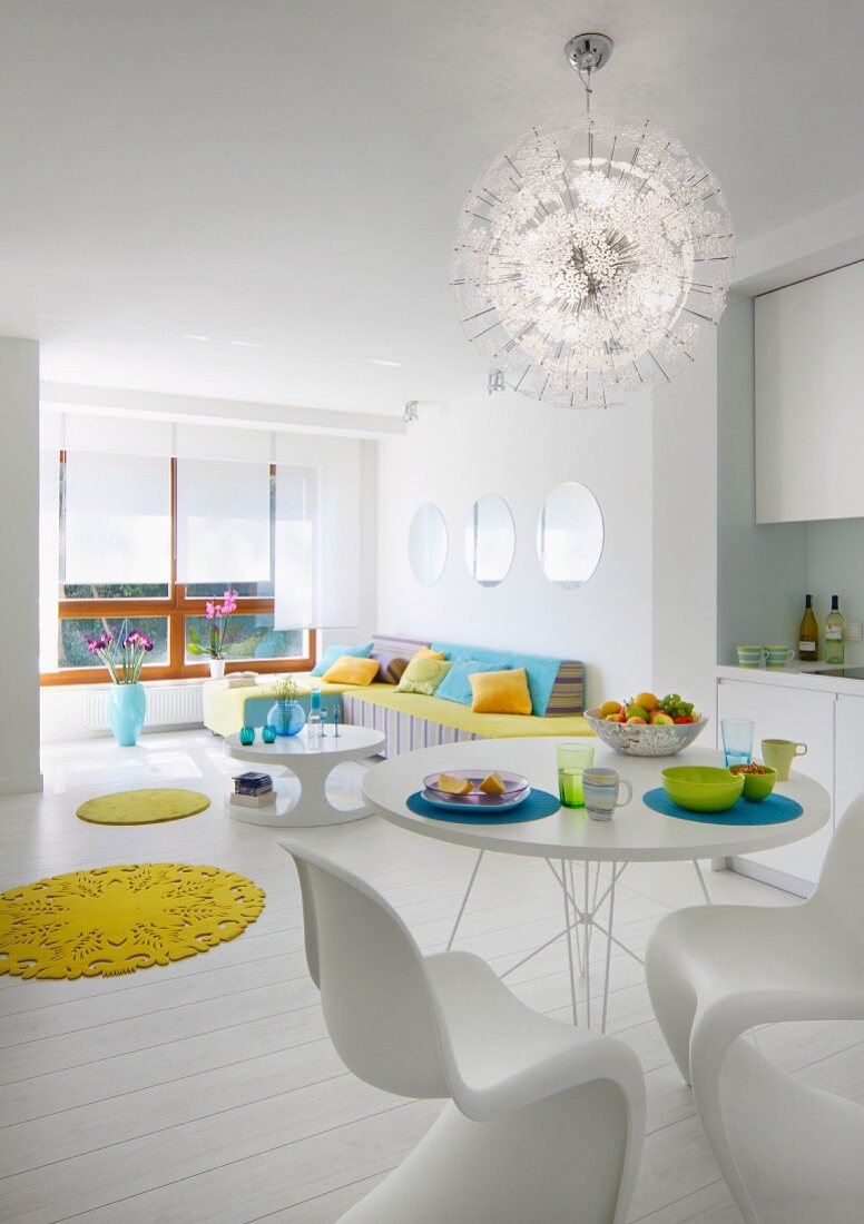 Offener Wohnraum in Weiß, Essplatz mit Klassiker Schalenstühlen unter strahlenförmiger Pendelleuchte, gelbe, runde Teppiche als Farbtupfer