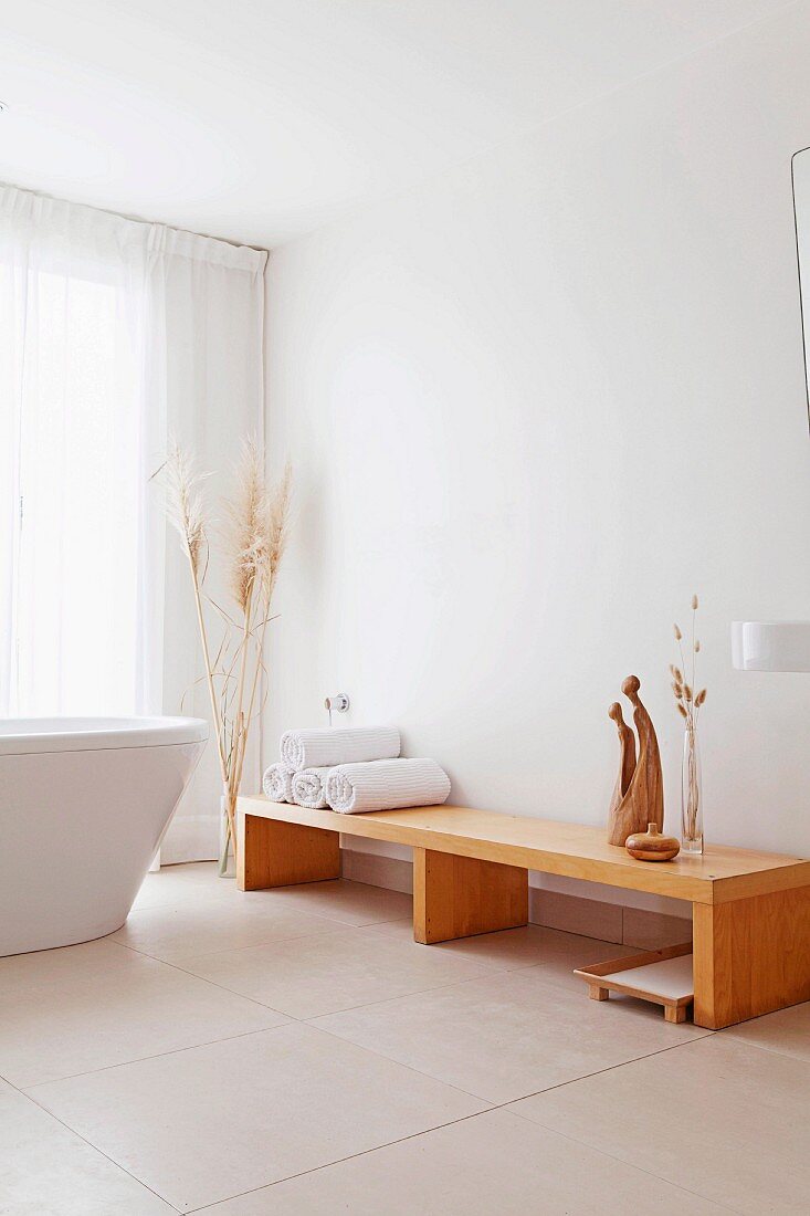 Rolled towels and sculptures on pale wooden bench, free-standing bathtub and large floor tiles in modern bathroom