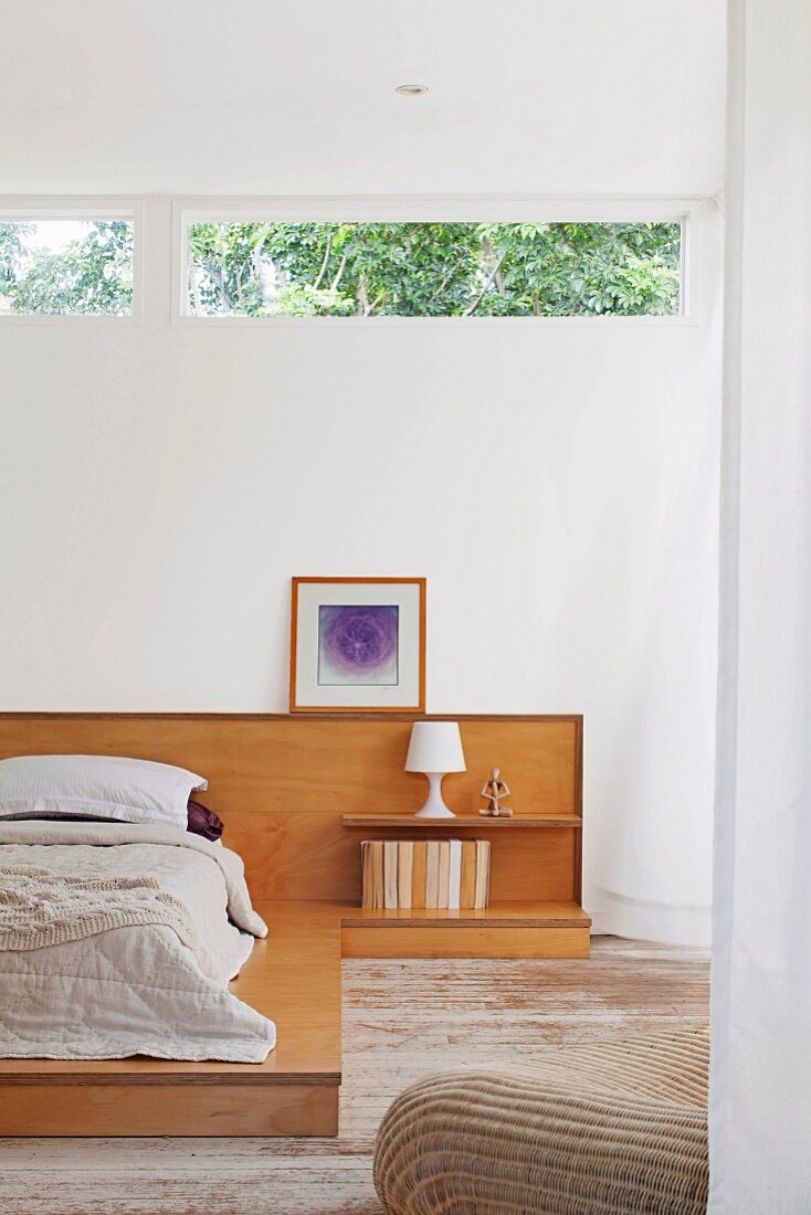 Bettlager auf Holzpodest mit halbhoher Holzverkleidung an Wand mit Oberlicht, seitlich Nachttischleuchte auf Ablage