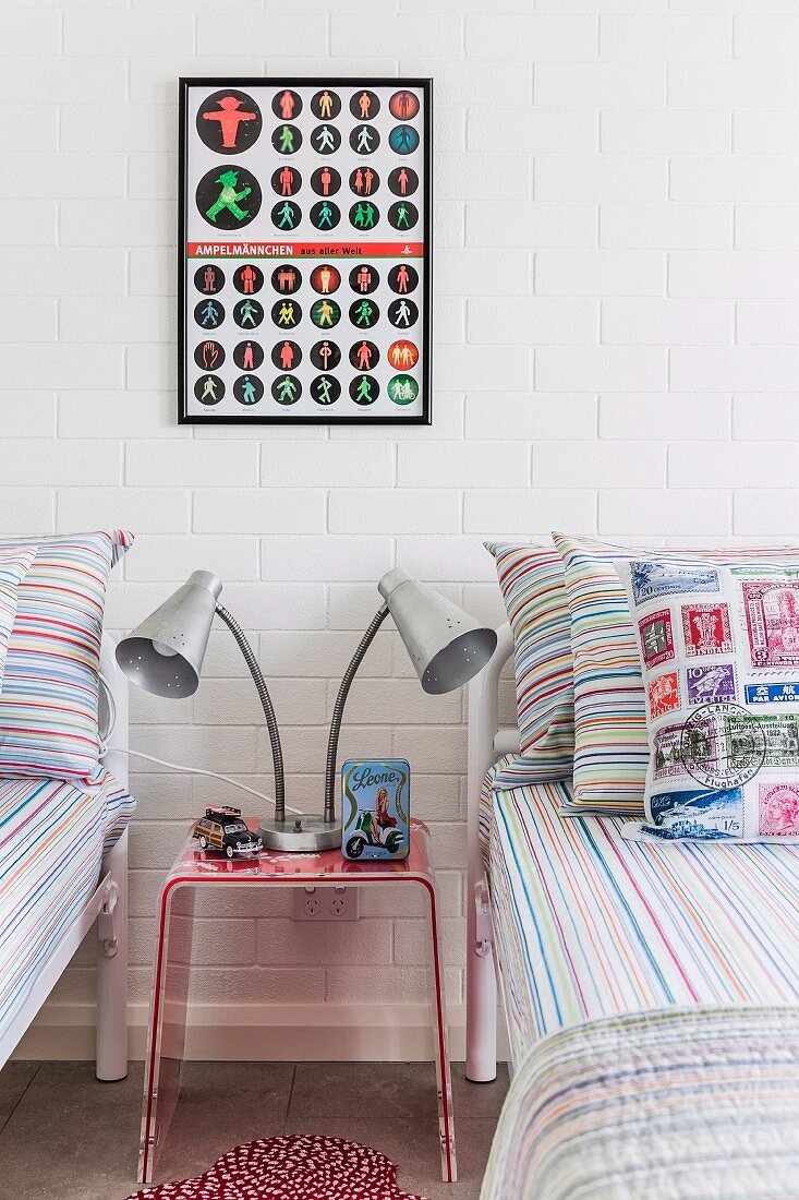 Table lamp with two heads on plexiglas bedside table between twin beds below framed poster on whitewashed brick wall