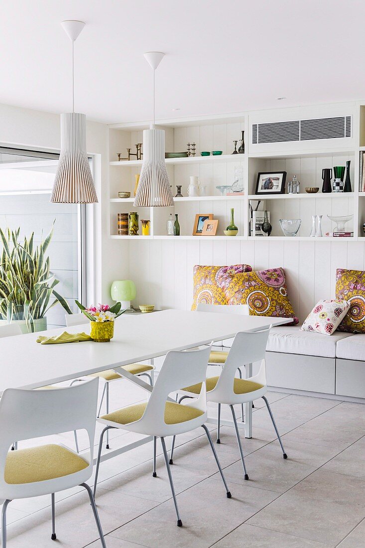 White dining table, chairs with seat cushions, pendant lamps with slatted lampshades and cushions on bench in background below wall-mounted shelves