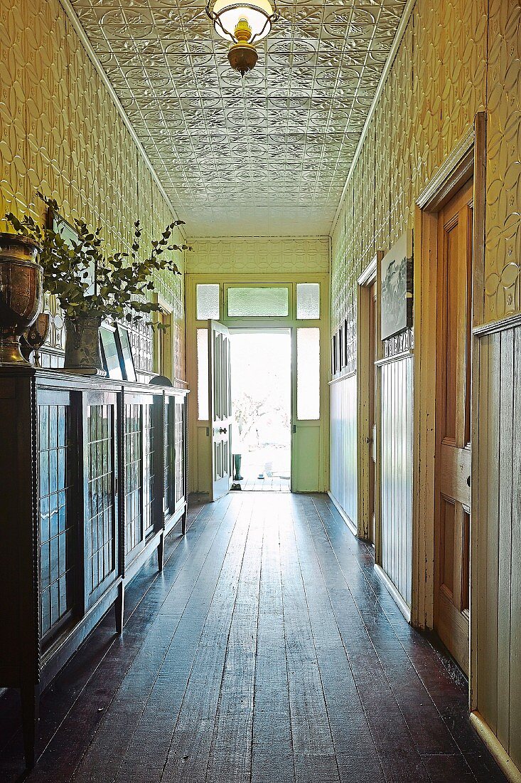 Schmaler, langer Flur mit antikem Sideboard auf Dielenboden, ornamentale Metallfliesen an Wand und Decke