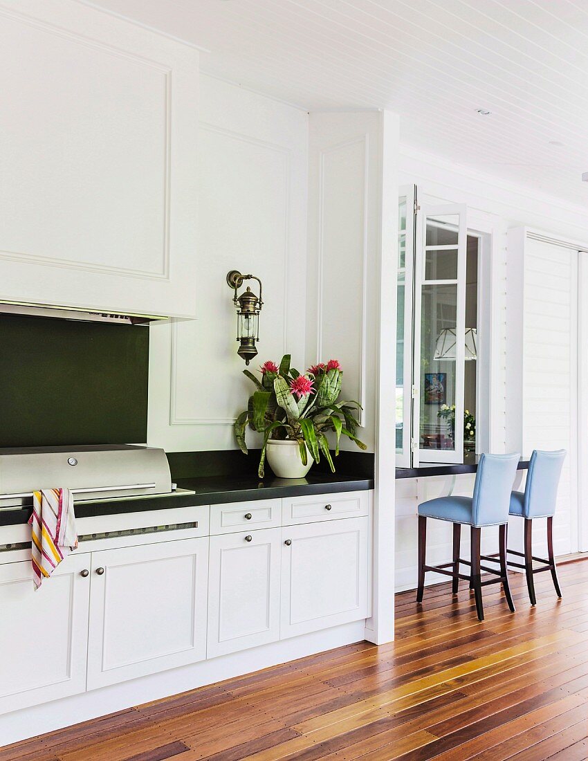 Kitchen counter with dark worksurface, white base units and seating area to one side on elegant veranda with wooden floor