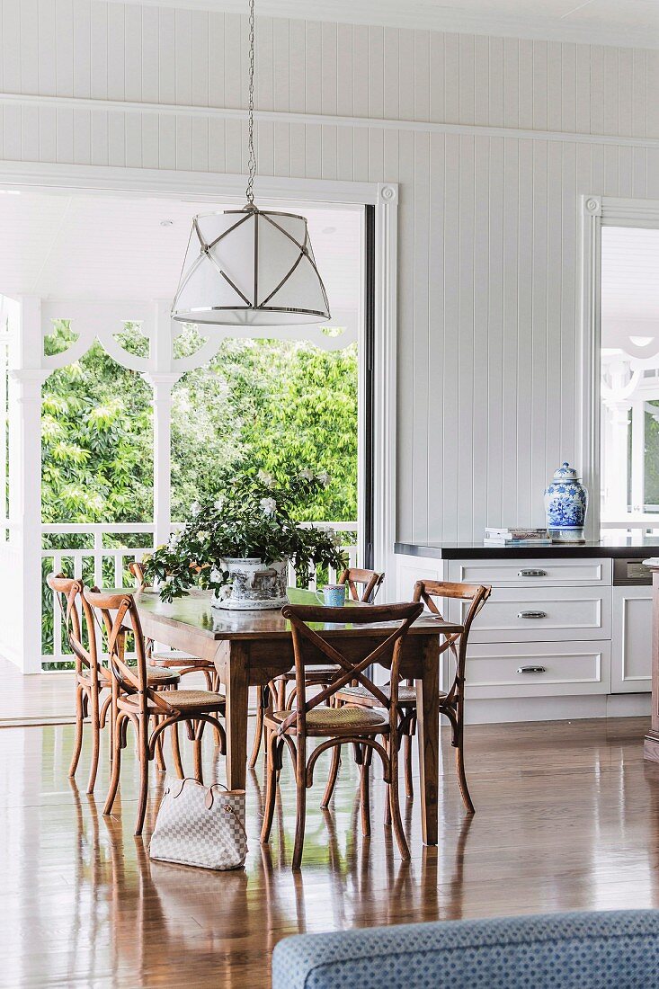 Ornate, Colonial-style wooden chairs below elegant pendant lamps in dining area in front of French windows